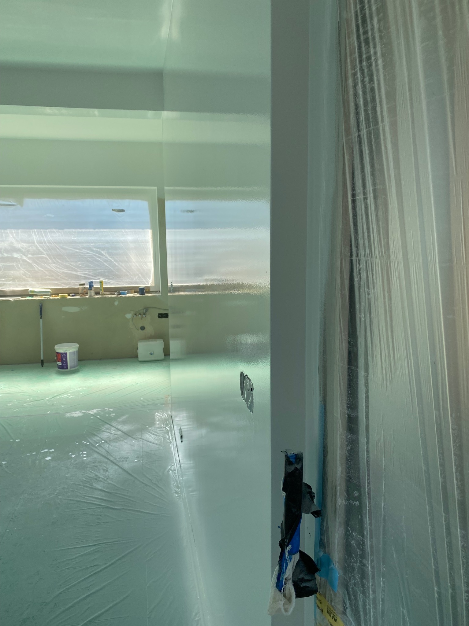 Świeżo pomalowane na biało wnętrze w Lubinie, zabezpieczone folią malarską. Widoczne narzędzia malarskie i wiadro z farbą przy oknie. Jasne, jednolite wykończenie.