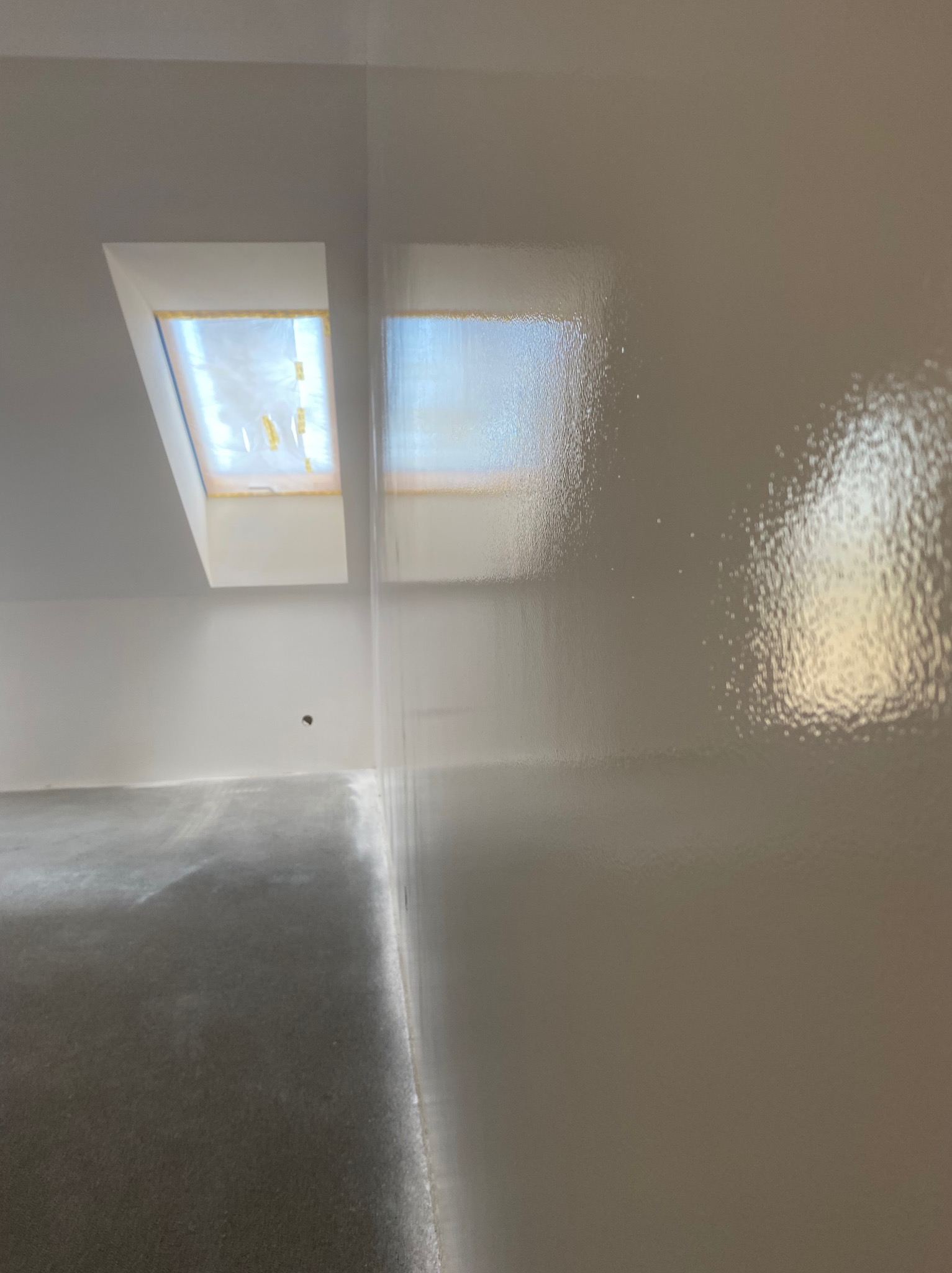 Świeżo pomalowana, gładka ściana w jasnym odcieniu z widocznym oknem dachowym. Podłoga w surowym stanie, przygotowana do dalszych prac wykończeniowych.