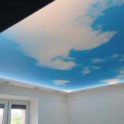 BUDiREM Darek Polaczek - Pokój z sufitem napinanym z motywem błękitnego nieba i białych chmur, oświetlonym listwą LED wokół obwodu, widok z dołu, białe ściany i okno po lewej stronie.