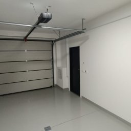 BUDiREM Darek Polaczek - Wykończone wnętrze garażu z automatyczną bramą segmentową, szarą posadzką żywiczną i białymi ścianami, widoczny napęd bramy pod sufitem oraz czarne drzwi wejściowe do garażu.