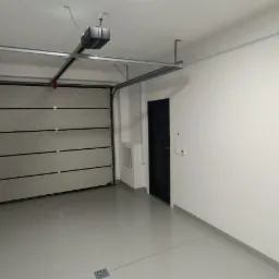 Wykończone wnętrze garażu z automatyczną bramą segmentową, szarą posadzką żywiczną i białymi ścianami, widoczny napęd bramy pod sufitem oraz czarne drzwi wejściowe do garażu.