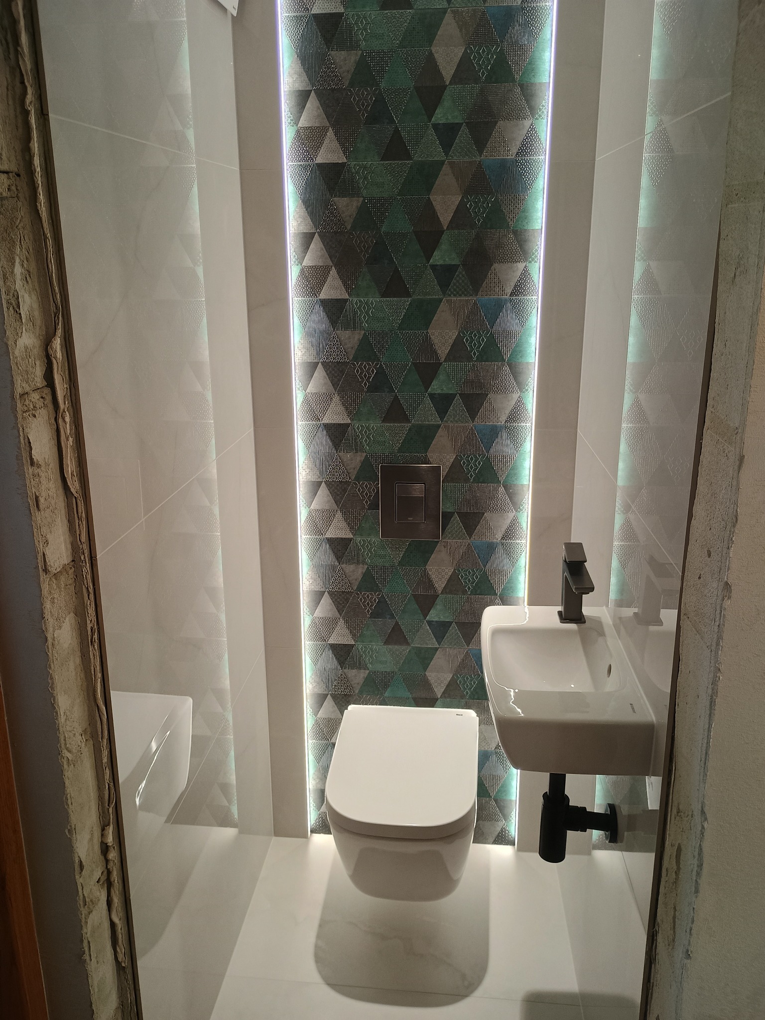 Nowoczesna łazienka z podświetlaną ścianą z płytek o geometrycznym wzorze, białą ceramiką i czarną armaturą, widok z korytarza w trakcie prac wykończeniowych.