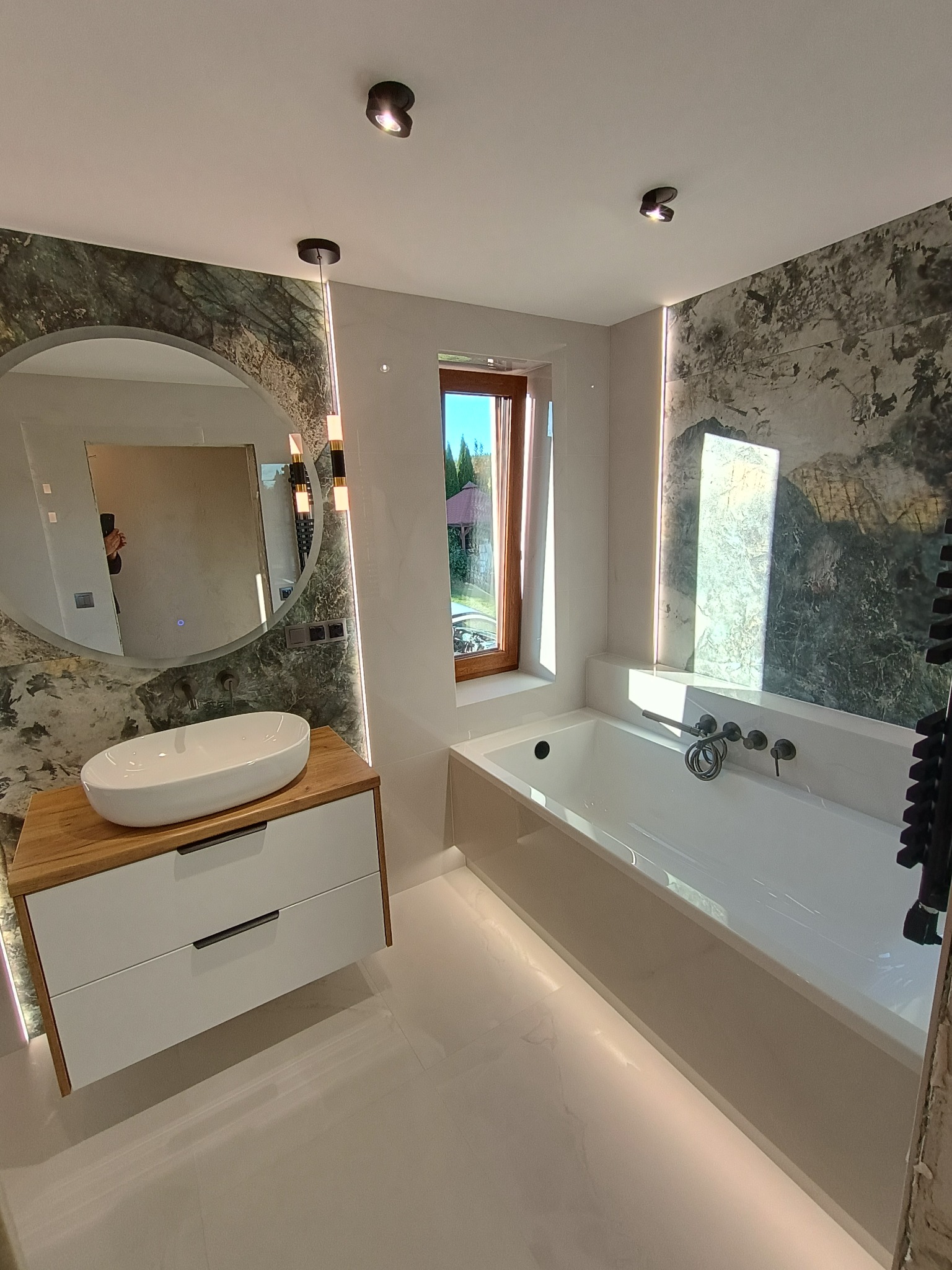 Nowoczesna łazienka z owalną umywalką nablatową na drewnianej szafce, okrągłym lustrem z podświetleniem, wanną i dekoracyjną okładziną ścienną imitującą kamień.