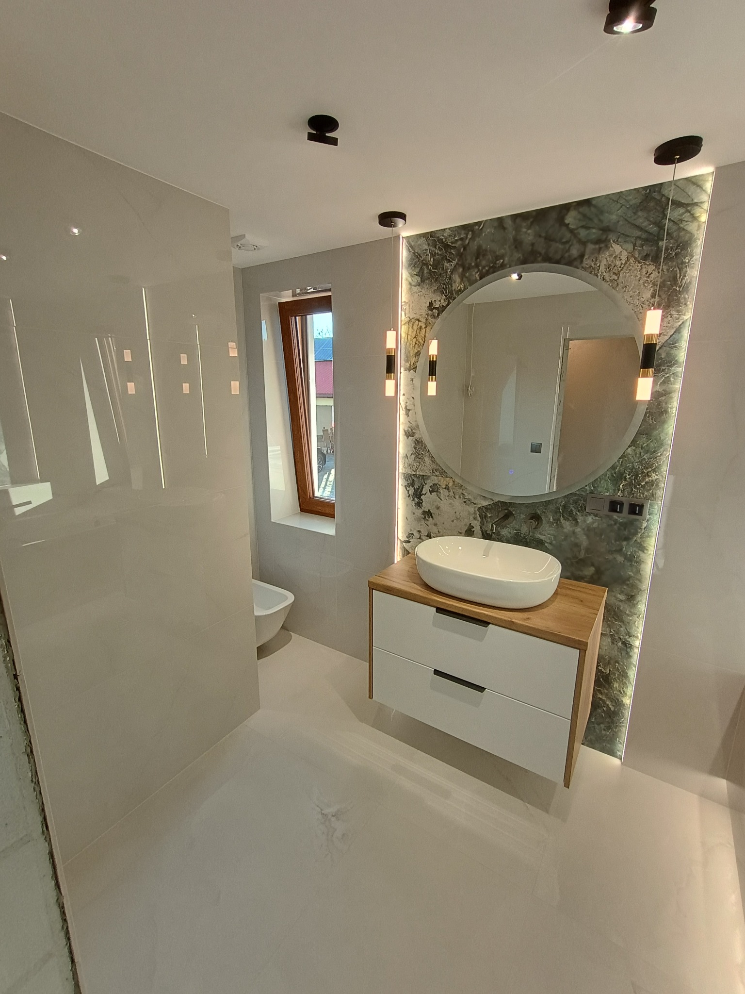Nowoczesna łazienka z owalną umywalką nablatową na szafce z drewnianym blatem i białymi frontami, okrągłe lustro z podświetleniem na ścianie z zielonego marmuru, jasne płytki na ścianach i podłodze...