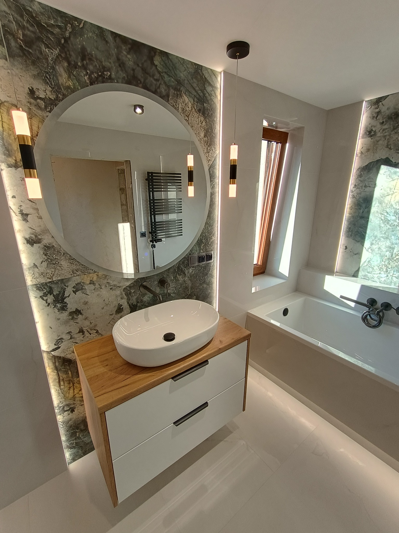 Nowoczesna łazienka z okrągłym lustrem podświetlonym LED, umywalką nablatową na szafce z drewnianym blatem i białymi frontami, oraz wanną pod skosem okna dachowego.
