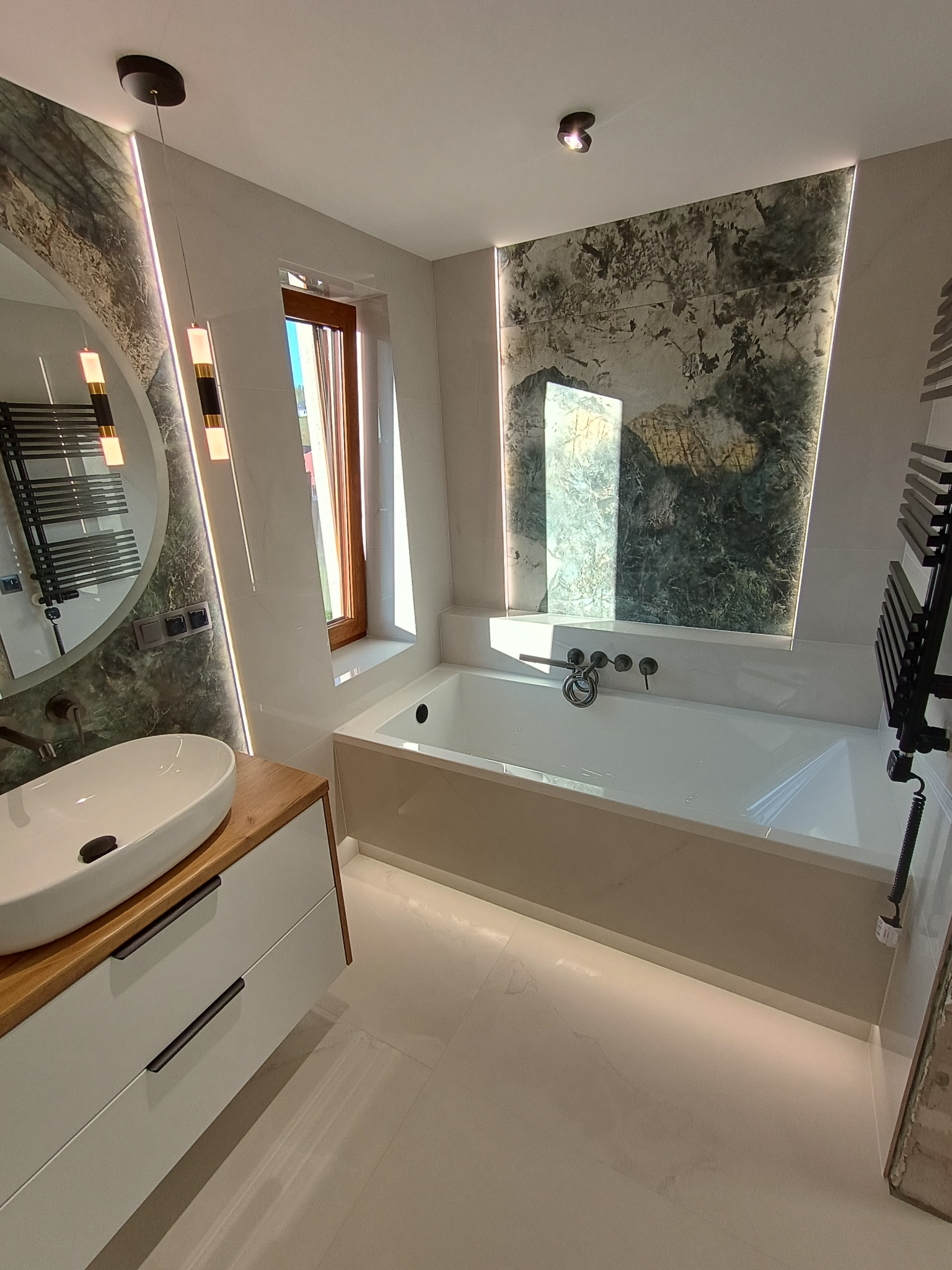 Nowoczesna łazienka z owalną umywalką nablatową na drewnianym blacie, białą wanną zabudowaną z mozaiką imitującą kamień na ścianie i czarnym grzejnikiem drabinkowym.