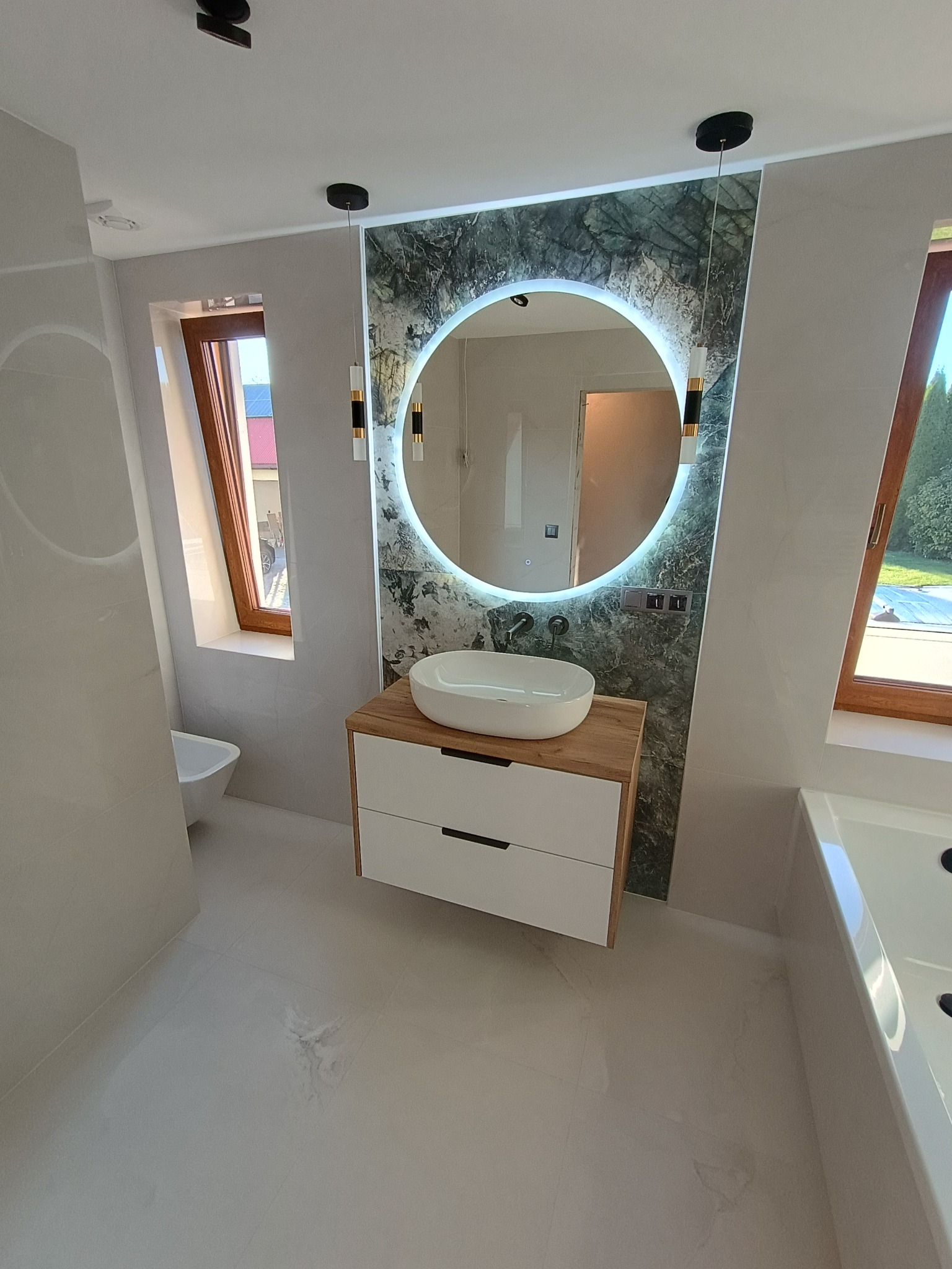 Nowoczesna łazienka z okrągłym lustrem podświetlanym LED, umywalką nablatową na szafce z drewnianym blatem i białymi frontami, płytki imitujące zielony marmur na ścianie, oraz widoczne okno dachowe.