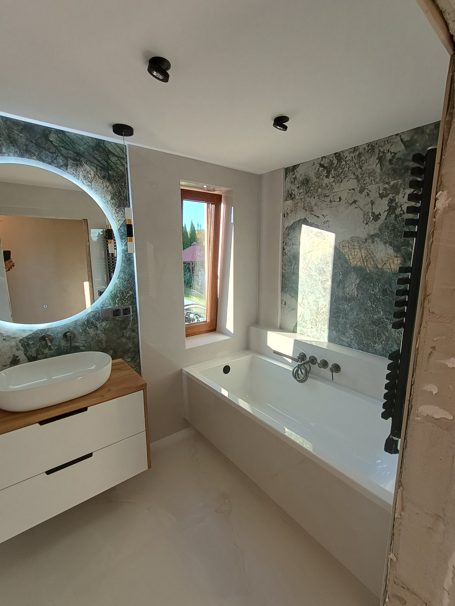 Nowoczesna łazienka w Dubiecku w trakcie remontu, z owalną umywalką na drewnianej szafce, podświetlanym lustrem, wanną i grzejnikiem drabinkowym, z widocznymi śladami prac wykończeniowych na ścianie.