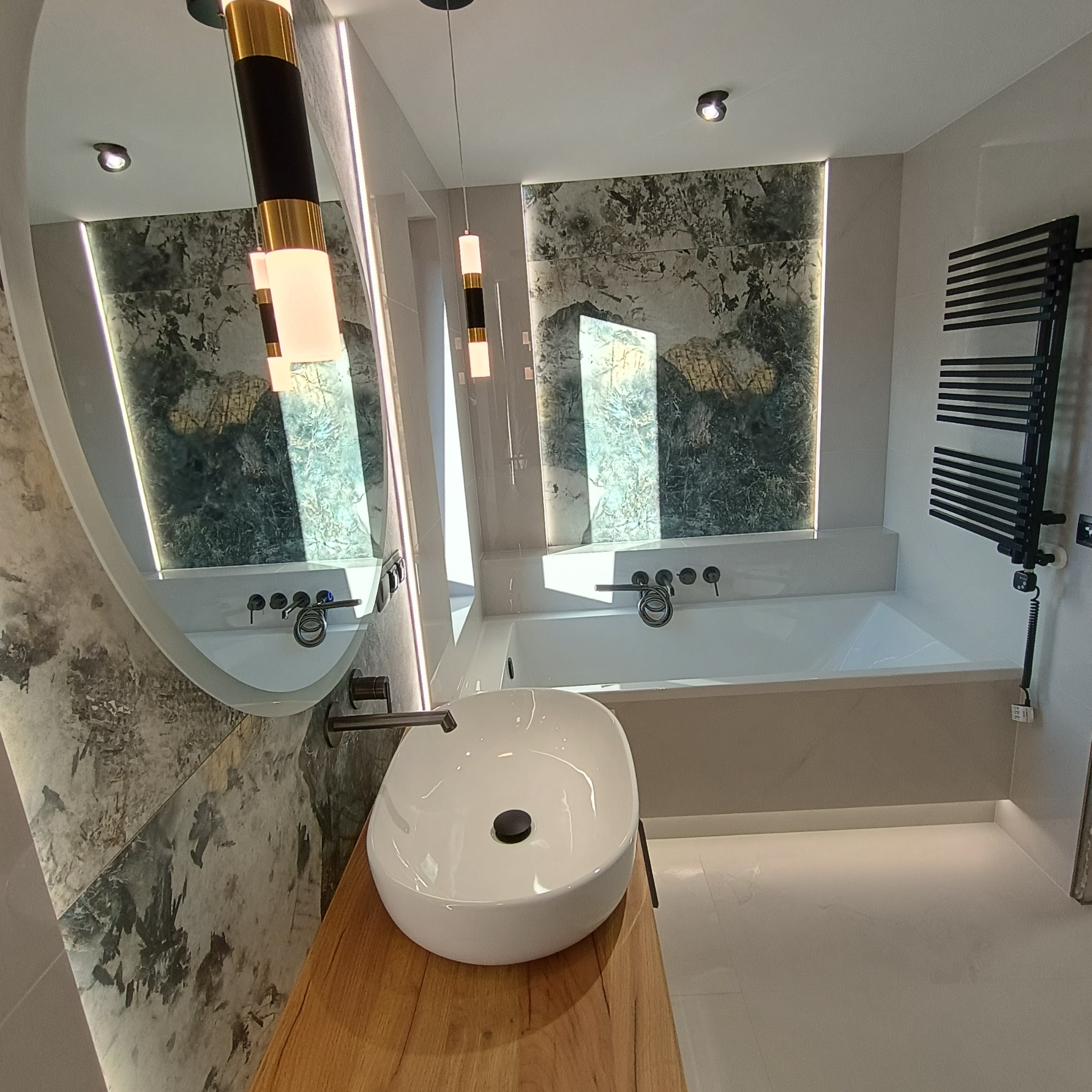 Nowoczesna łazienka z owalnym lustrem z podświetleniem LED, umywalką nablatową na drewnianym blacie, wanną oraz grzejnikiem drabinkowym w kolorze czarnym.