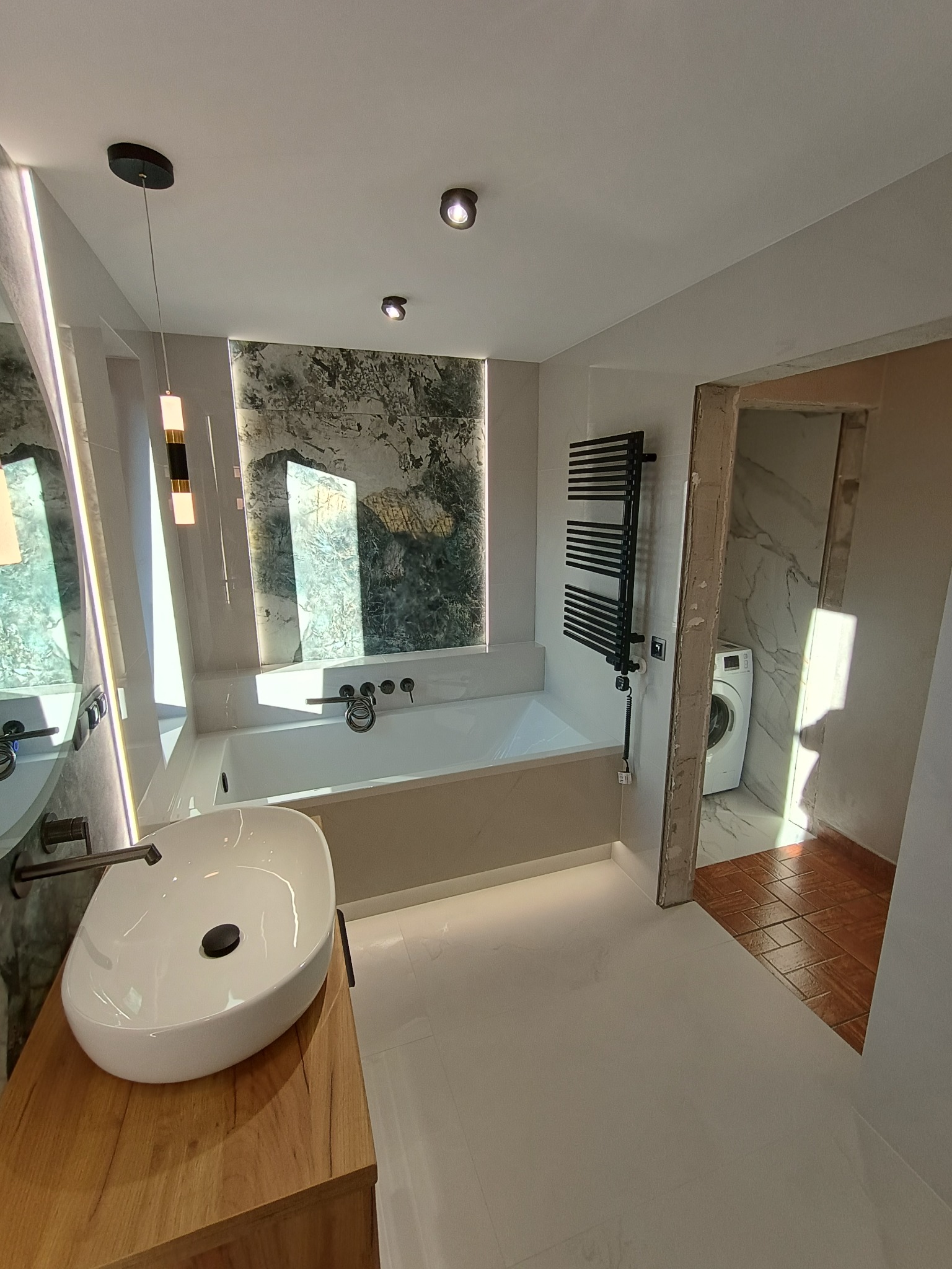 Nowoczesna łazienka z wolnostojącą umywalką na drewnianej szafce, białą wanną pod oknem z marmurowym wzorem, grzejnikiem drabinkowym i pralką widoczną w sąsiednim pomieszczeniu.