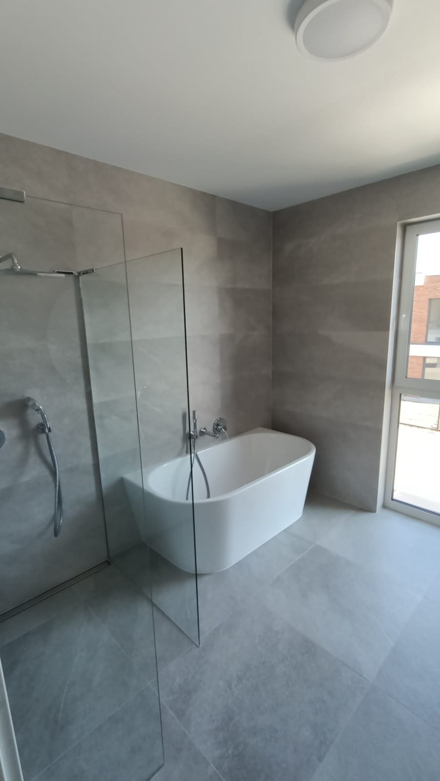 Nowoczesna łazienka z wolnostojącą białą wanną, szklaną kabiną prysznicową i szarymi płytkami w formacie 60x60, oświetlona okrągłą lampą sufitową.