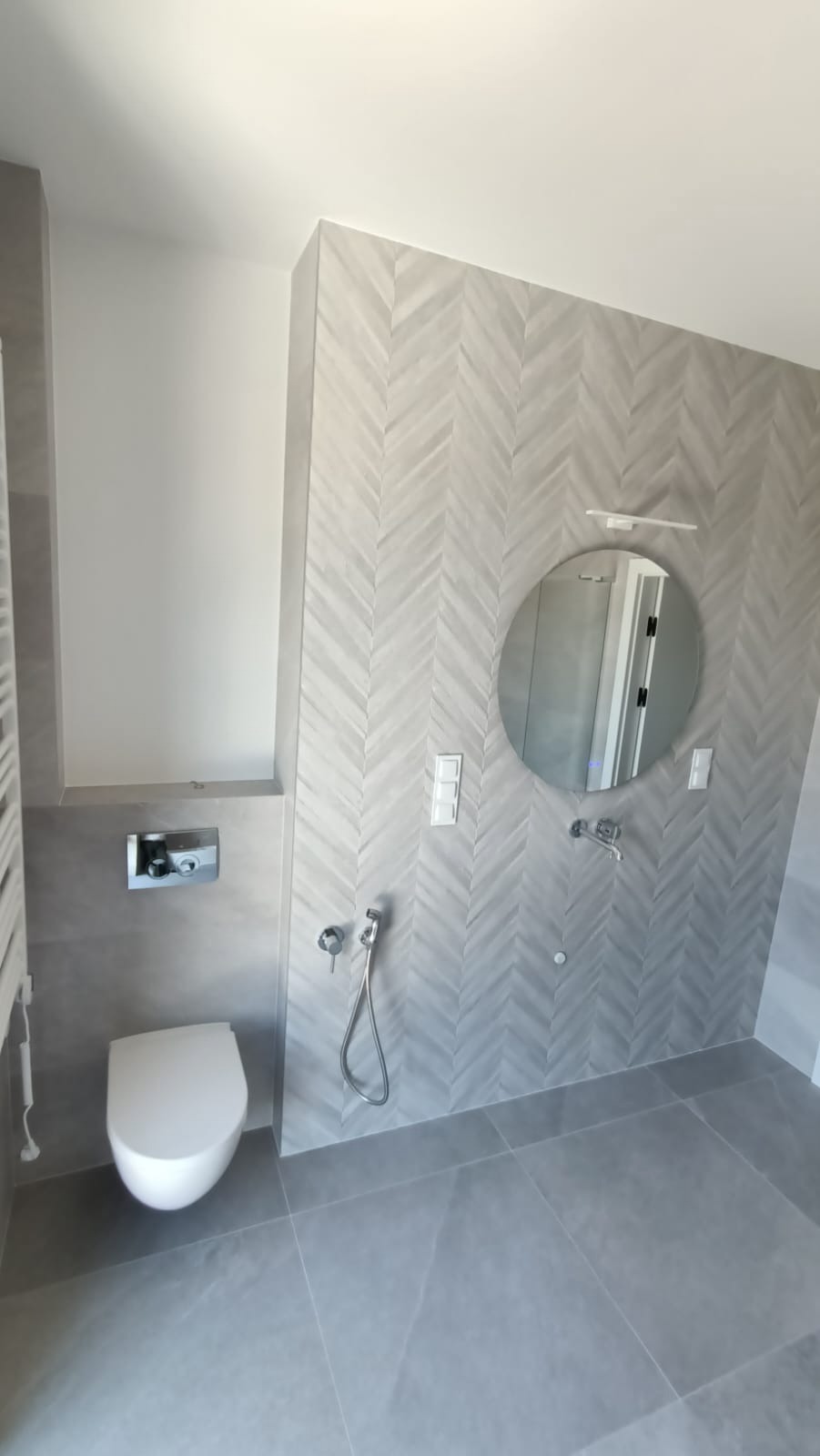 Nowoczesna łazienka w odcieniach szarości z płytkami ułożonymi w jodełkę na ścianie, okrągłe lustro z podświetleniem i wisząca toaleta.