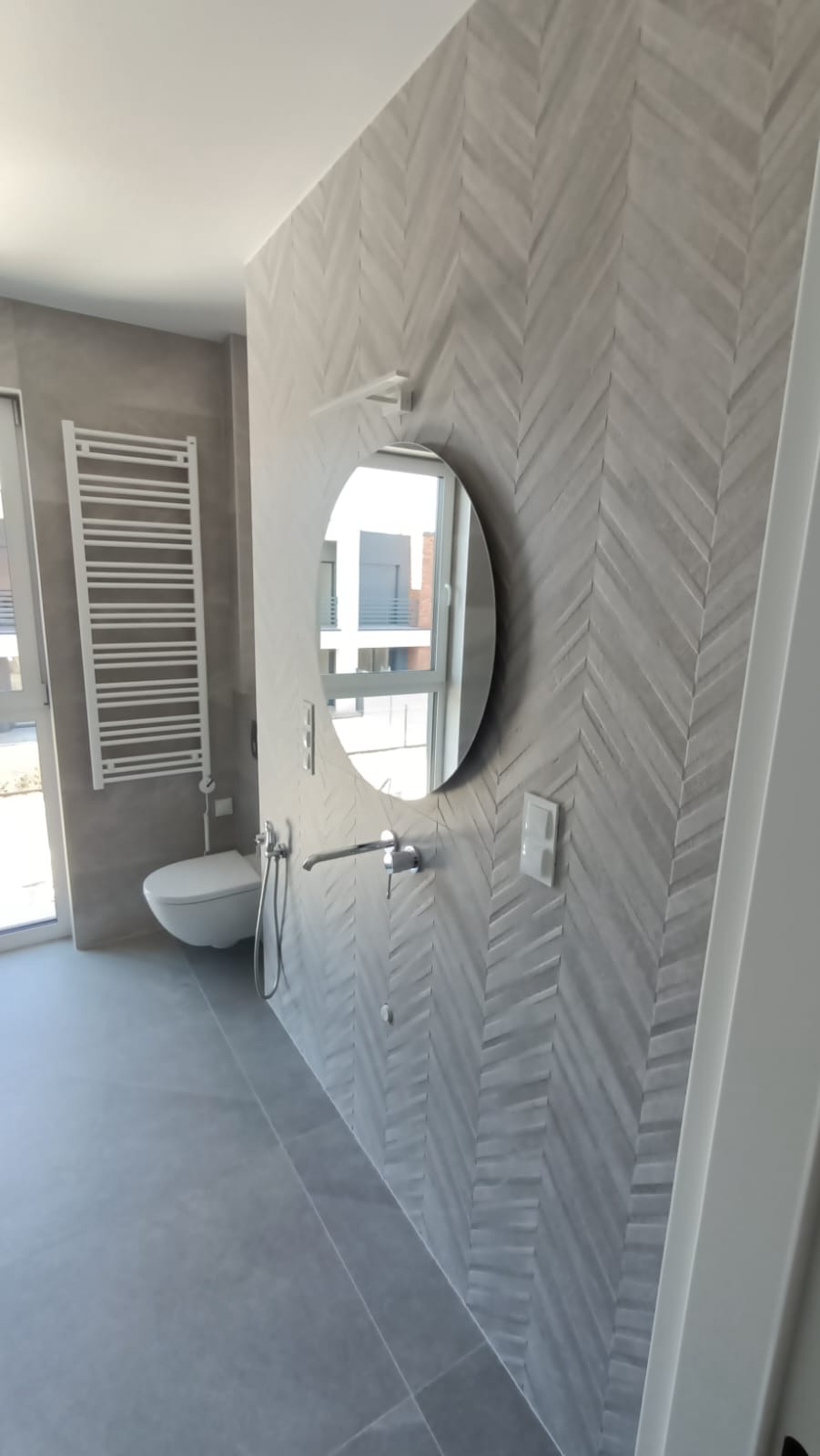 Nowoczesna łazienka w odcieniach szarości z płytkami ułożonymi w jodełkę na ścianie, owalne lustro z chromowaną baterią, biały grzejnik drabinkowy i podwieszana toaleta.