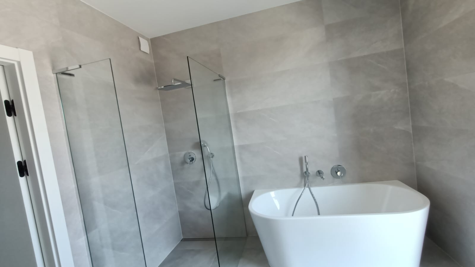 Nowoczesna łazienka w odcieniach szarości z wolnostojącą białą wanną i przeszkloną kabiną prysznicową typu walk-in z deszczownicą.