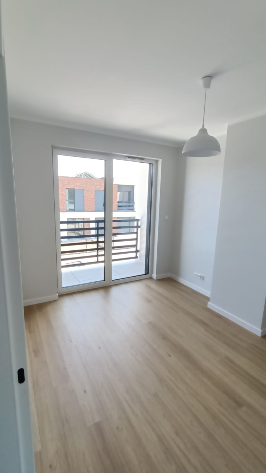 Jasne wnętrze pokoju z panelami podłogowymi w odcieniach drewna, białe ściany i sufit z wiszącą lampą, okno balkonowe z widokiem na sąsiedni budynek z balkonami.