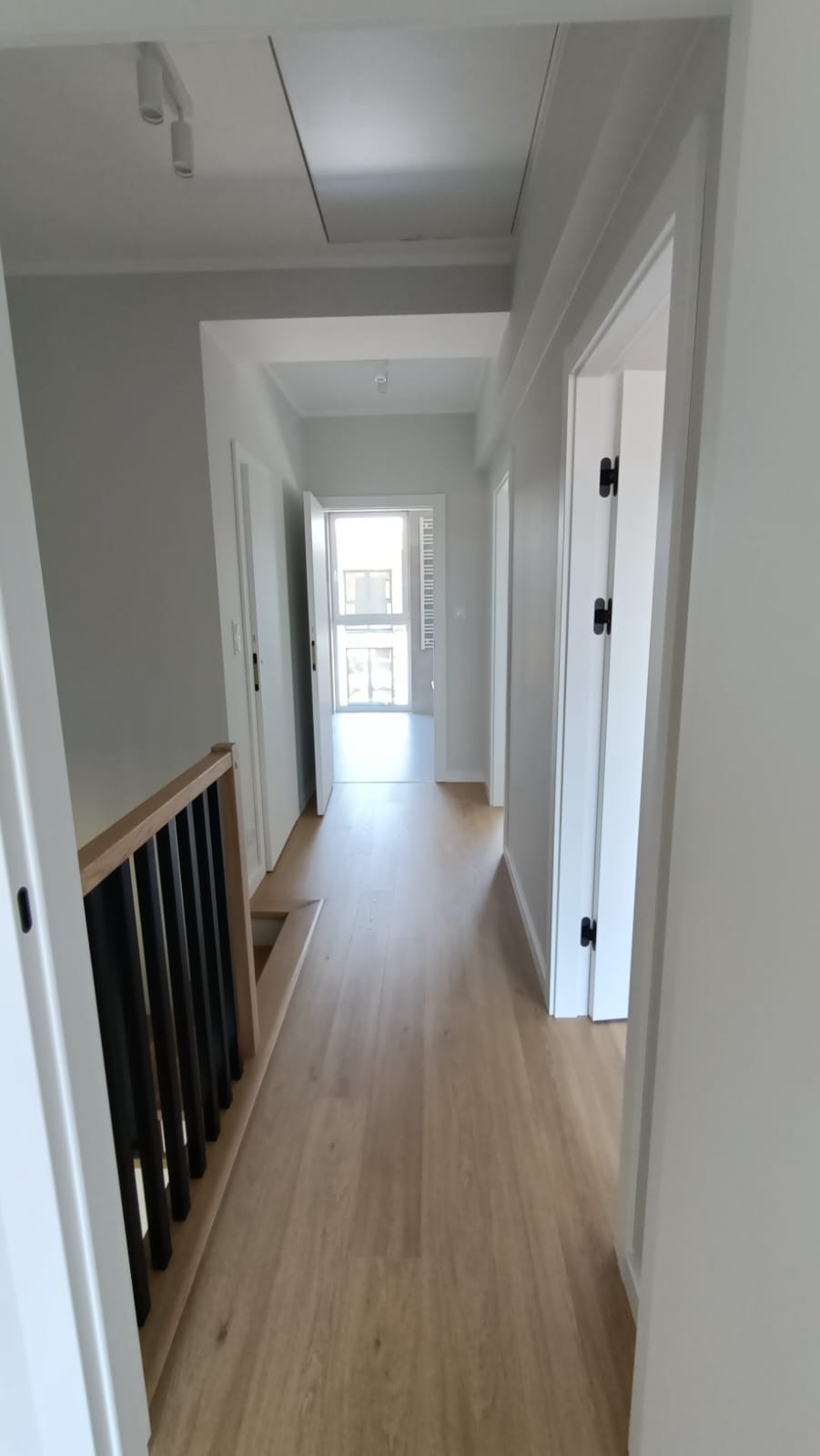 Długi korytarz w jasnych odcieniach szarości i beżu, z drewnianą podłogą i czarną balustradą schodów, drzwi z czarnymi klamkami po obu stronach, w głębi widoczne okno.