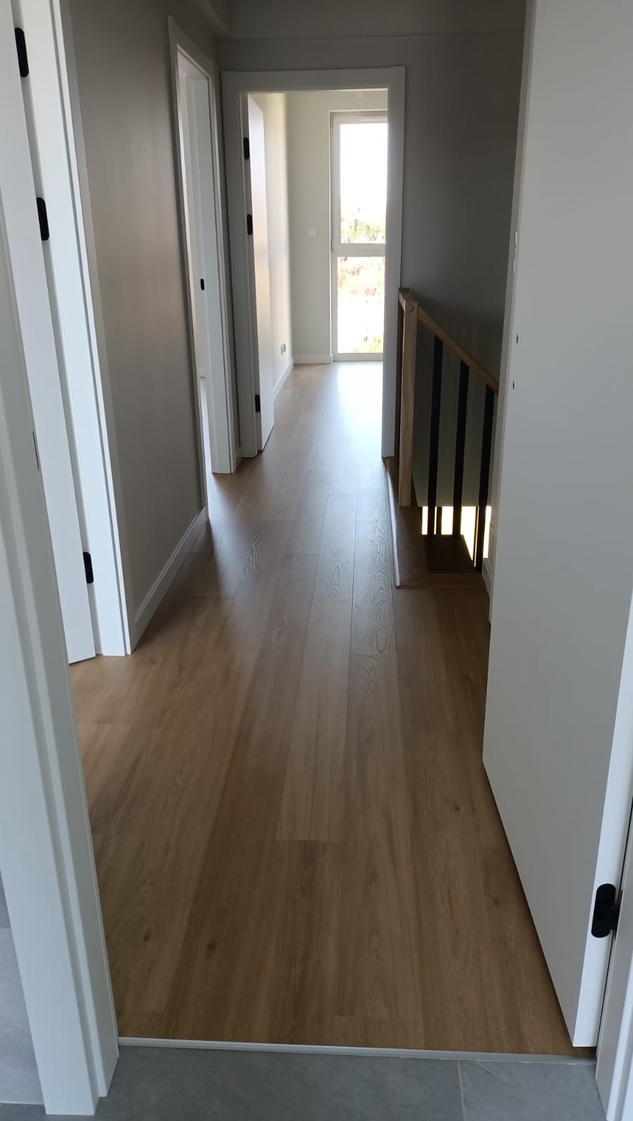 Korytarz z nową drewnopodobną podłogą, widoczne białe drzwi z czarnymi klamkami i fragment schodów z drewnianą balustradą w jasnym wnętrzu.