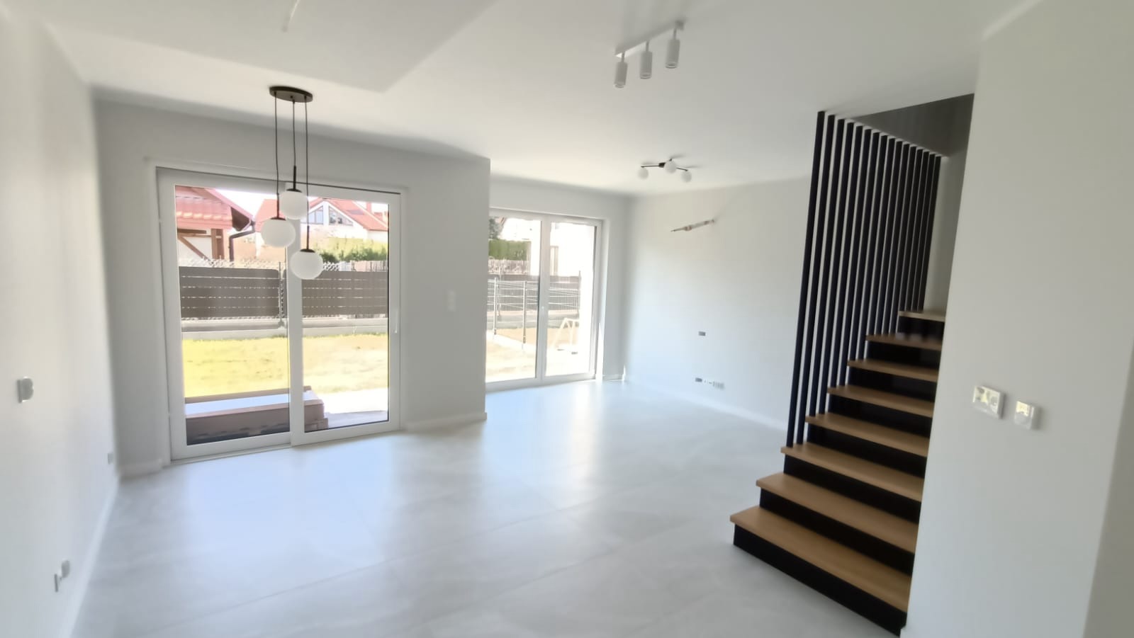 Jasne, przestronne wnętrze domu po remoncie, z widocznymi schodami z drewnianymi stopniami i czarnymi elementami, białymi ścianami i podłogą, oraz oknami wychodzącymi na ogród.