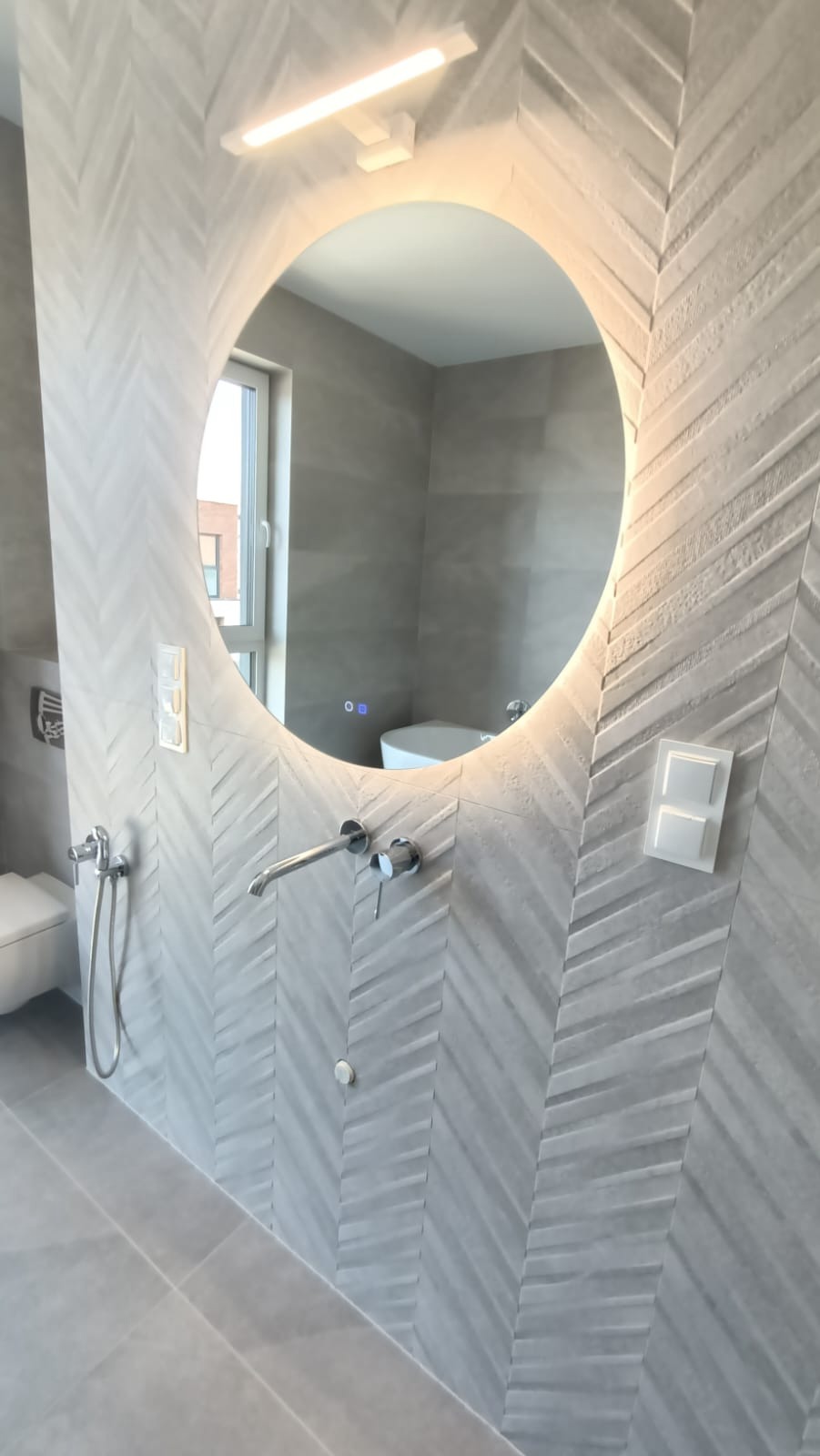 Nowoczesna łazienka z okrągłym lustrem podświetlanym LED, ściany wyłożone szarą płytką ułożoną we wzór jodełki, widoczna bateria umywalkowa i włącznik światła.