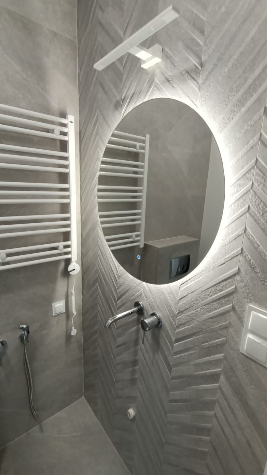 Szara łazienka z okrągłym lustrem z podświetleniem LED, grzejnikiem drabinkowym, baterią podtynkową i płytkami ułożonymi w jodełkę.