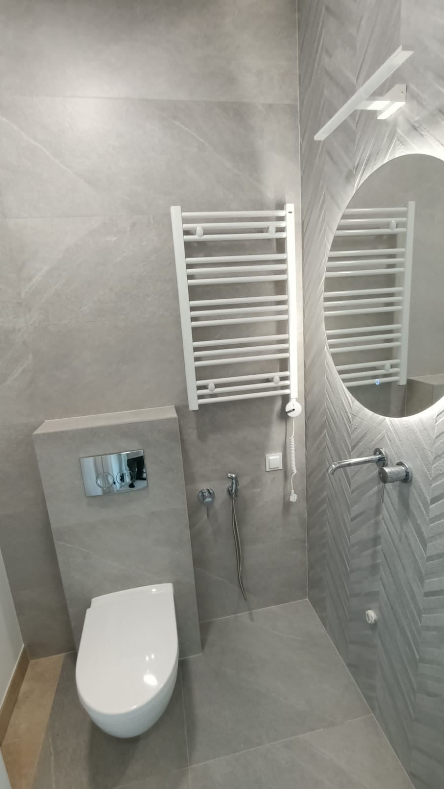 Nowoczesna łazienka w odcieniach szarości z podwieszaną toaletą, grzejnikiem drabinkowym i okrągłym lustrem z podświetleniem LED.