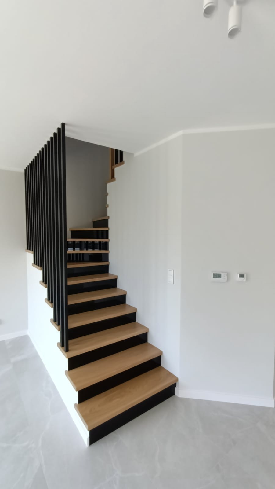 Nowoczesne schody drewniane z czarnymi podstopnicami i minimalistyczną balustradą z pionowych czarnych prętów w jasnym wnętrzu z szarą podłogą i białymi ścianami.