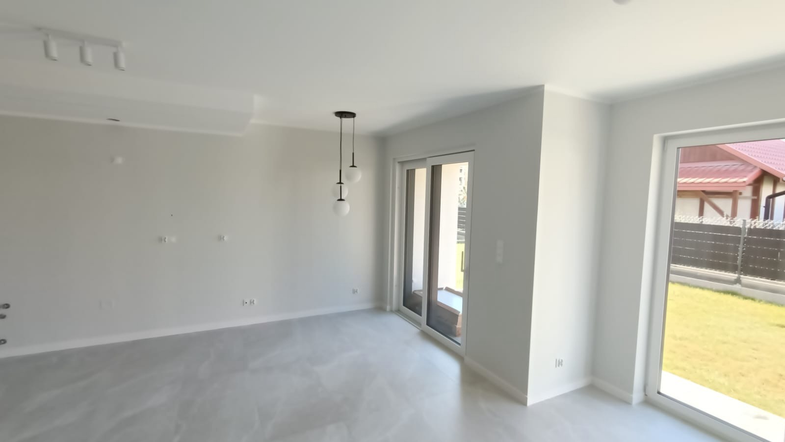 Jasne, przestronne wnętrze z białymi ścianami, szarą podłogą imitującą marmur, nowoczesną lampą wiszącą z trzema kulami i widokiem na ogród przez duże okno tarasowe.