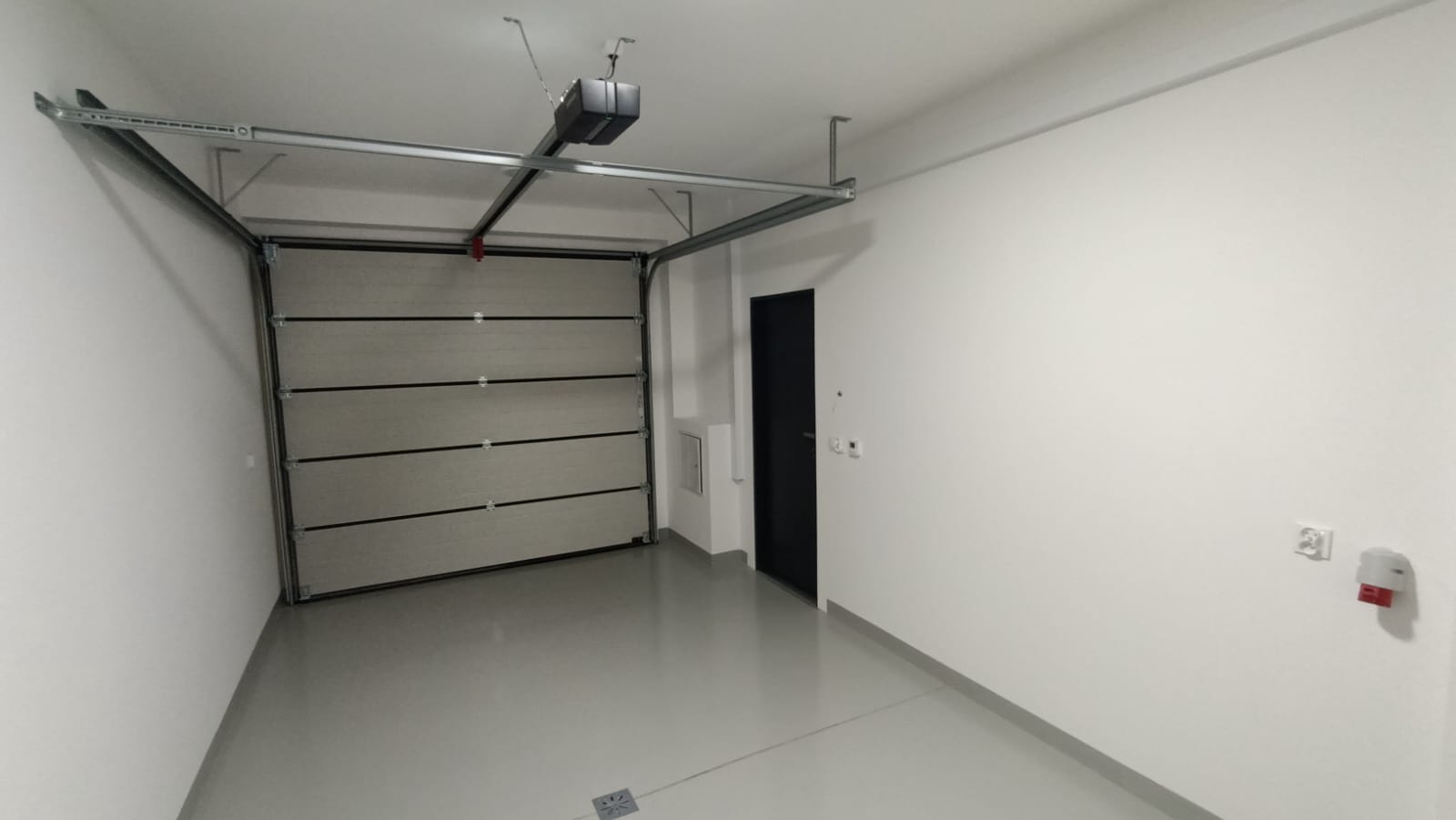 Wykończone wnętrze garażu z automatyczną bramą segmentową, szarą posadzką żywiczną i białymi ścianami, widoczny napęd bramy pod sufitem oraz czarne drzwi wejściowe do garażu.