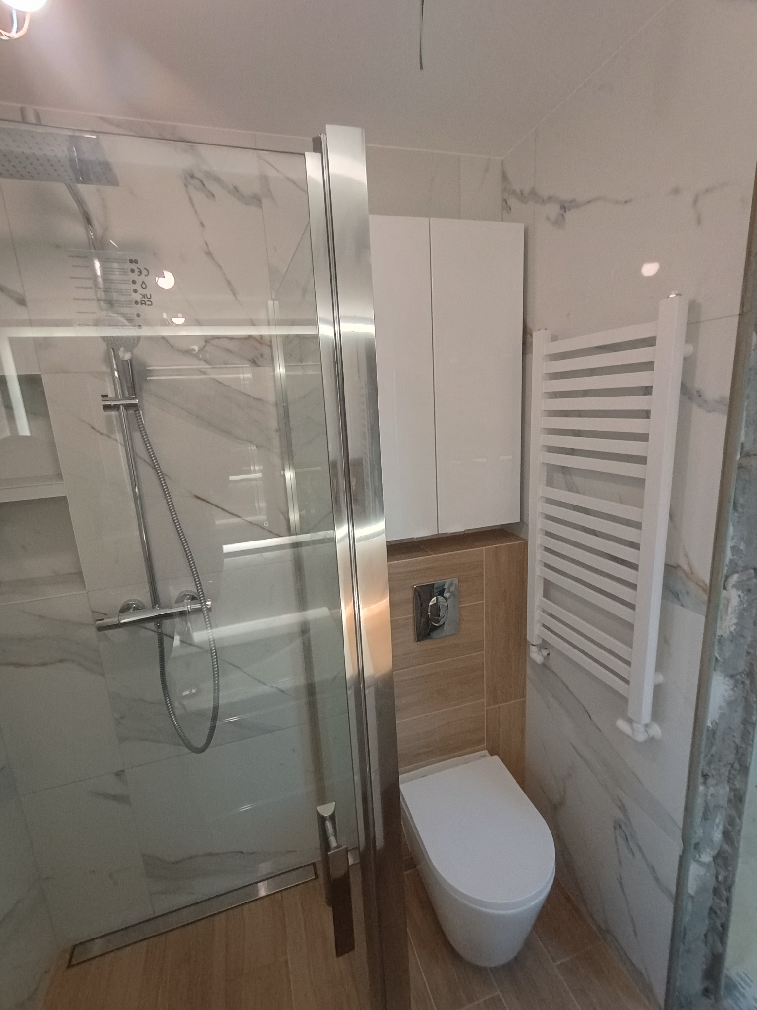 Nowoczesna łazienka z kabiną prysznicową typu walk-in, wiszącą toaletą, grzejnikiem drabinkowym i szafką nad zabudową WC, wykończona płytkami imitującymi marmur i drewno.