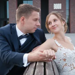 Uśmiechnięta panna młoda w koronkowej sukni spogląda na pana młodego w granatowym garniturze z muszką, siedząc na drewnianej ławce na tle nowoczesnego budynku z czerwonej cegły.