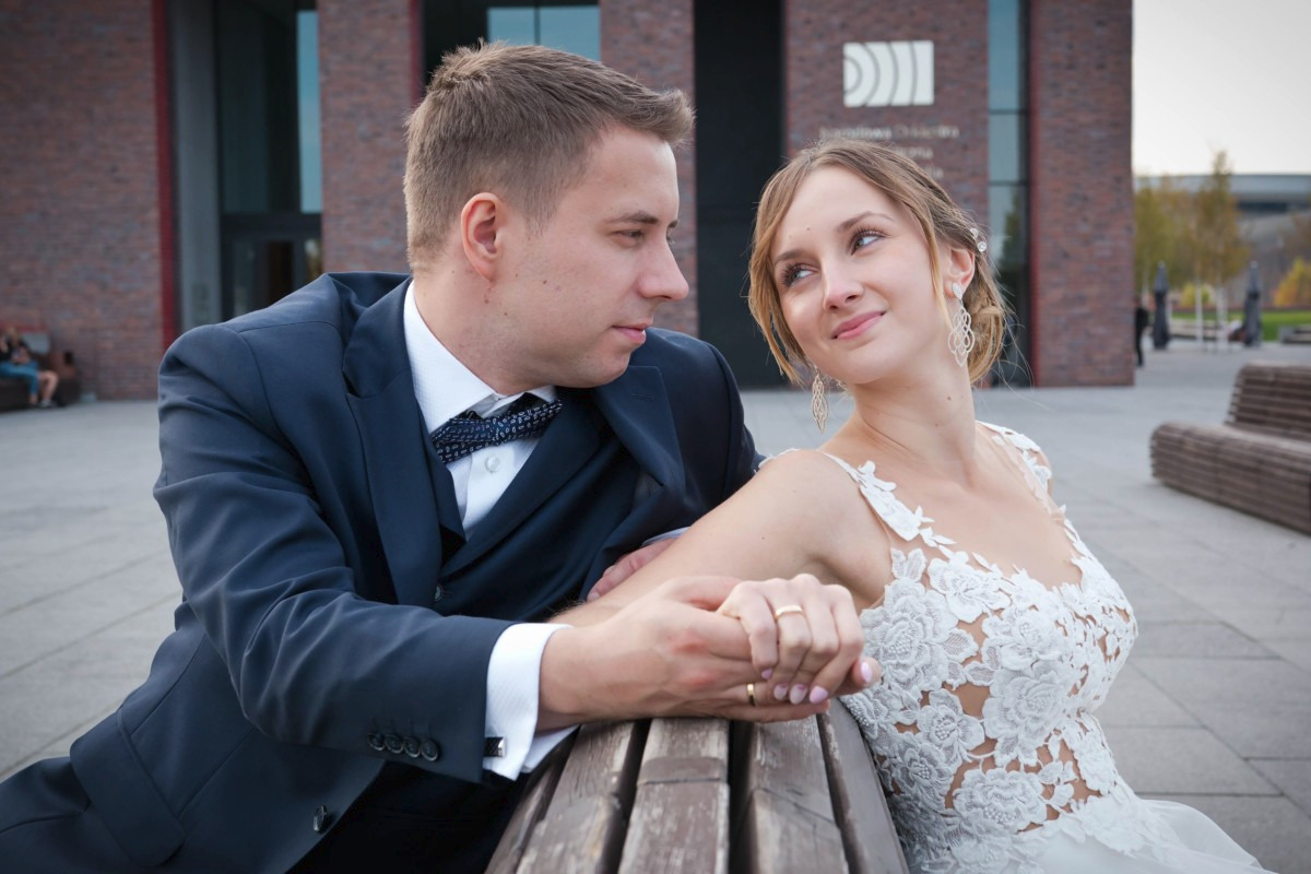 Uśmiechnięta panna młoda w koronkowej sukni spogląda na pana młodego w granatowym garniturze z muszką, siedząc na drewnianej ławce na tle nowoczesnego budynku z czerwonej cegły.