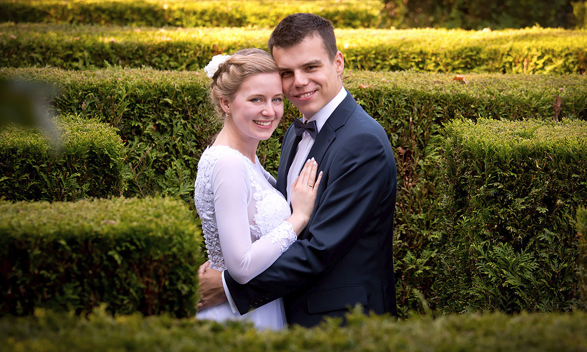 Uśmiechnięta para młoda w objęciach na tle geometrycznych żywopłotów; panna młoda w koronkowej sukni z długim rękawem, pan młody w granatowym garniturze z muchą w kropki.