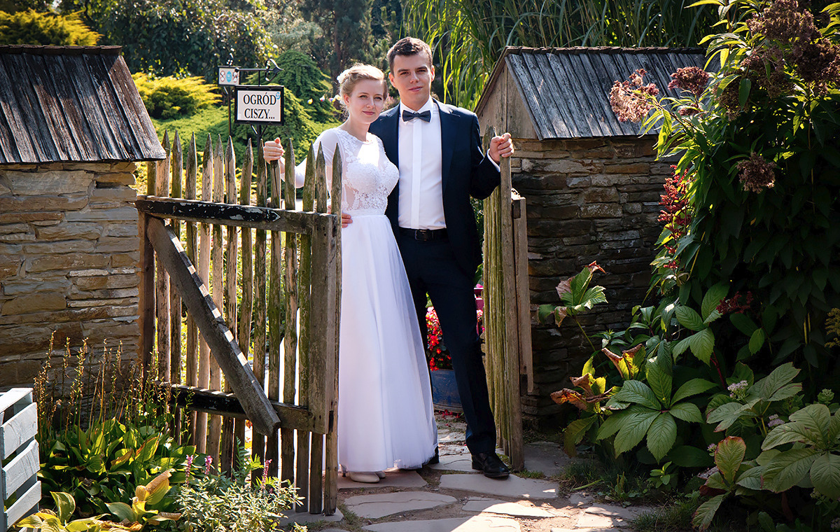 Para młoda pozuje przy drewnianej furtce w rustykalnym ogrodzie z kamiennymi budynkami w tle, tabliczka z napisem 'Ogród Ciszy'.