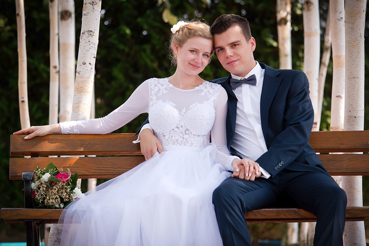 Para młoda siedząca na drewnianej ławce w parku, panna młoda w białej sukni z koronkową górą i długimi rękawami, pan młody w granatowym garniturze i muszce w kropki, bukiet ślubny leżący obok,...