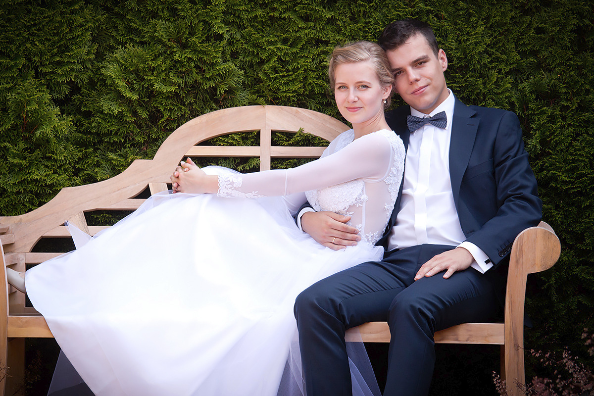 Para młoda siedzi na drewnianej ławce przed zielonym żywopłotem. Panna młoda w białej sukni z koronkowymi rękawami, pan młody w granatowym garniturze z muchą w kropki.