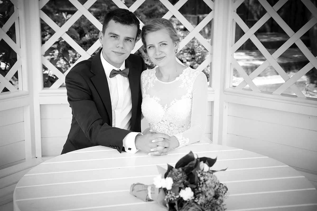 Czarno-białe zdjęcie pary młodej siedzącej przy okrągłym stole w altanie, panna młoda w koronkowej sukni z długimi rękawami, pan młody w garniturze z muszką, na stole leży bukiet.