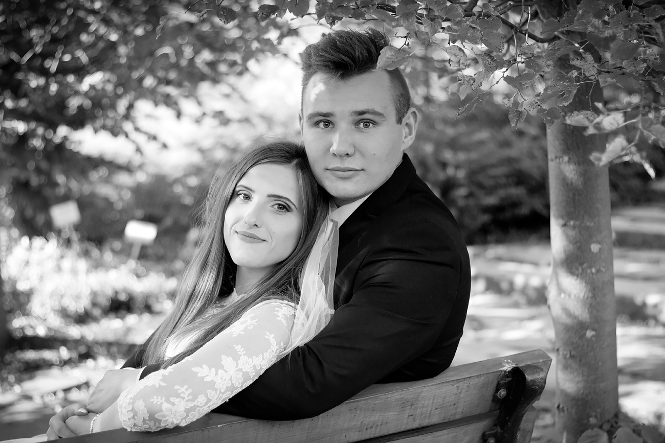 Czarno-białe portretowe ujęcie pary: kobieta w koronkowej sukience z długimi rękawami i mężczyzna w ciemnym garniturze, siedzący na ławce pod drzewem z liśćmi, delikatny uśmiech obojga.