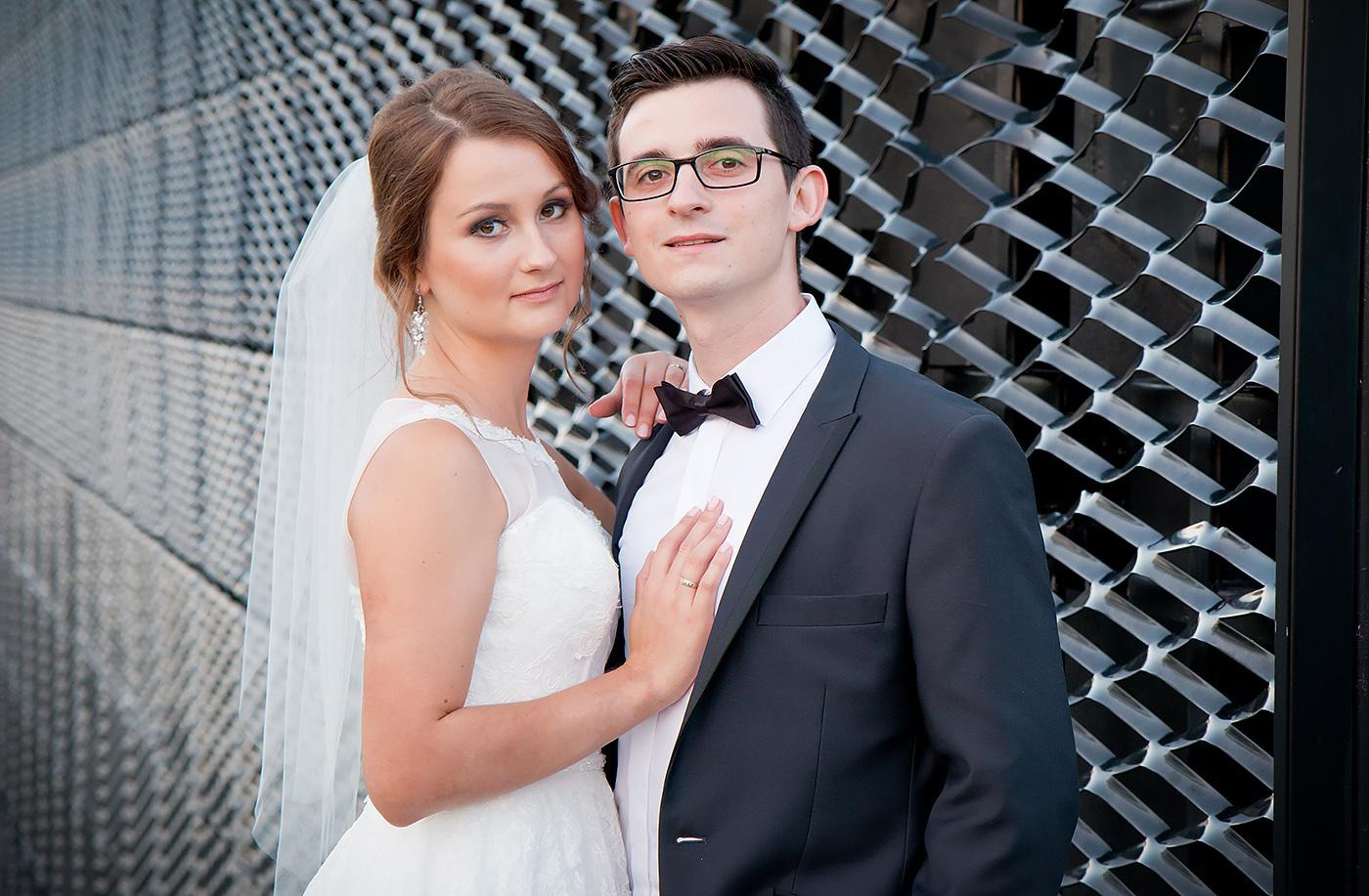 Para młoda w eleganckich strojach pozuje na tle nowoczesnej, metalowej konstrukcji z efektem 3D.