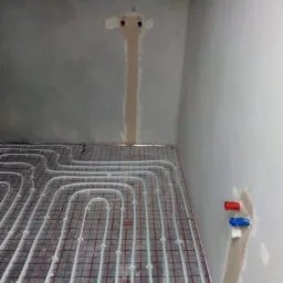 Białe rurki ogrzewania podłogowego ułożone na siatce z czerwonymi kwadratami, widoczne przyłącza w ścianie, surowy stan ścian, przygotowanie do wylewki.