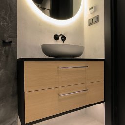 Krzysztof Murzynowski - Nowoczesna łazienka z wiszącą szafką z dwoma szufladami o jasnym, drewnianym froncie, grafitową umywalką nablatową i okrągłym lustrem z podświetleniem LED. Widoczne czarne elementy armatury...