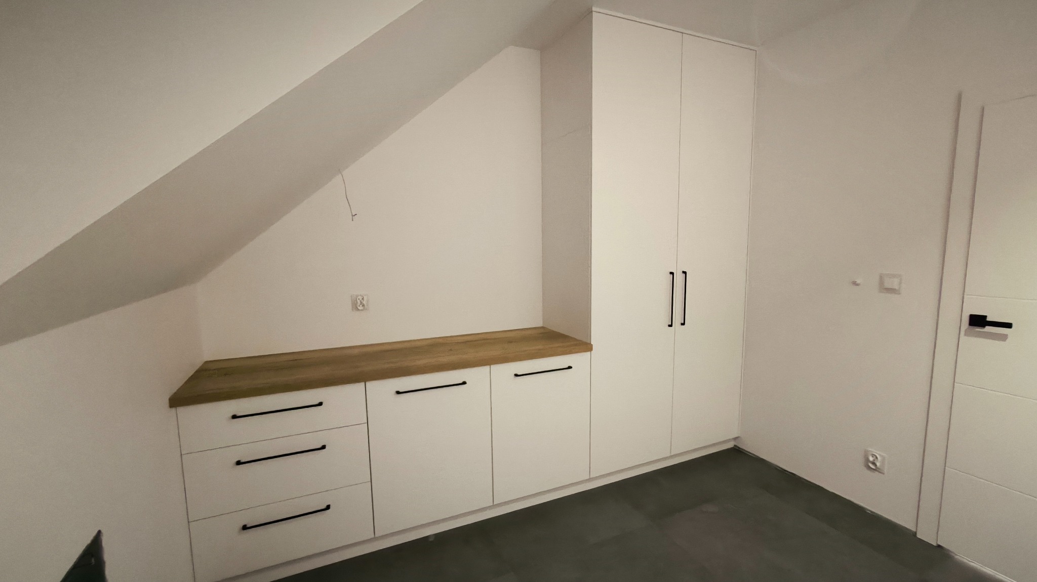 Biała szafa z szufladami i blatem z drewna wkomponowana w skos poddasza, czarne uchwyty, szara podłoga.