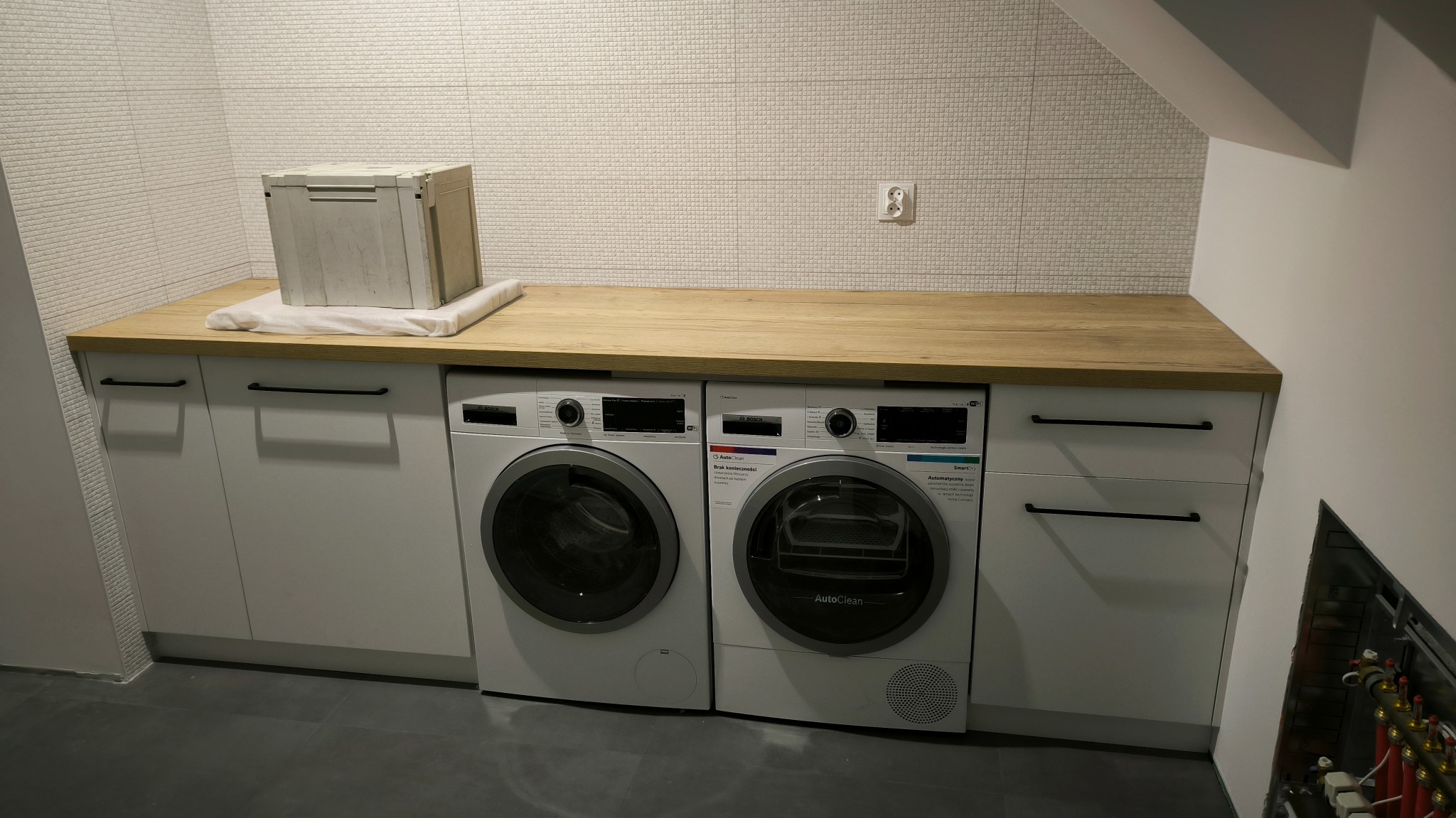 Nowoczesna pralnia z białymi szafkami, drewnianym blatem, pralką i suszarką Bosch Serie 8, białymi płytkami na ścianie i szarą podłogą, z skrzynią narzędziową na blacie.