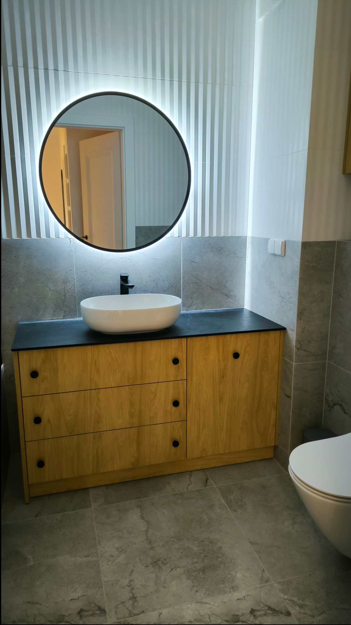 Nowoczesna łazienka z drewnianą szafką pod umywalkę z czarnym blatem i okrągłym lustrem podświetlanym LED. Na ścianie tapeta w pionowe pasy i szare płytki imitujące marmur.