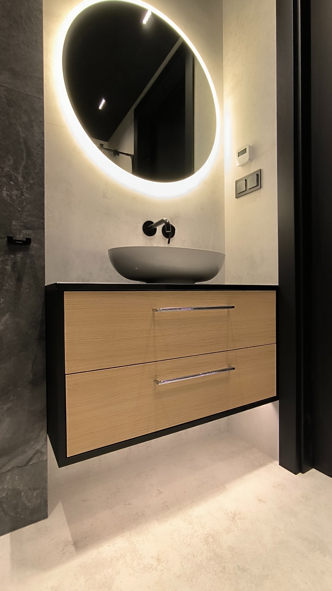 Nowoczesna łazienka z wiszącą szafką z dwoma szufladami o jasnym, drewnianym froncie, grafitową umywalką nablatową i okrągłym lustrem z podświetleniem LED. Widoczne czarne elementy armatury...