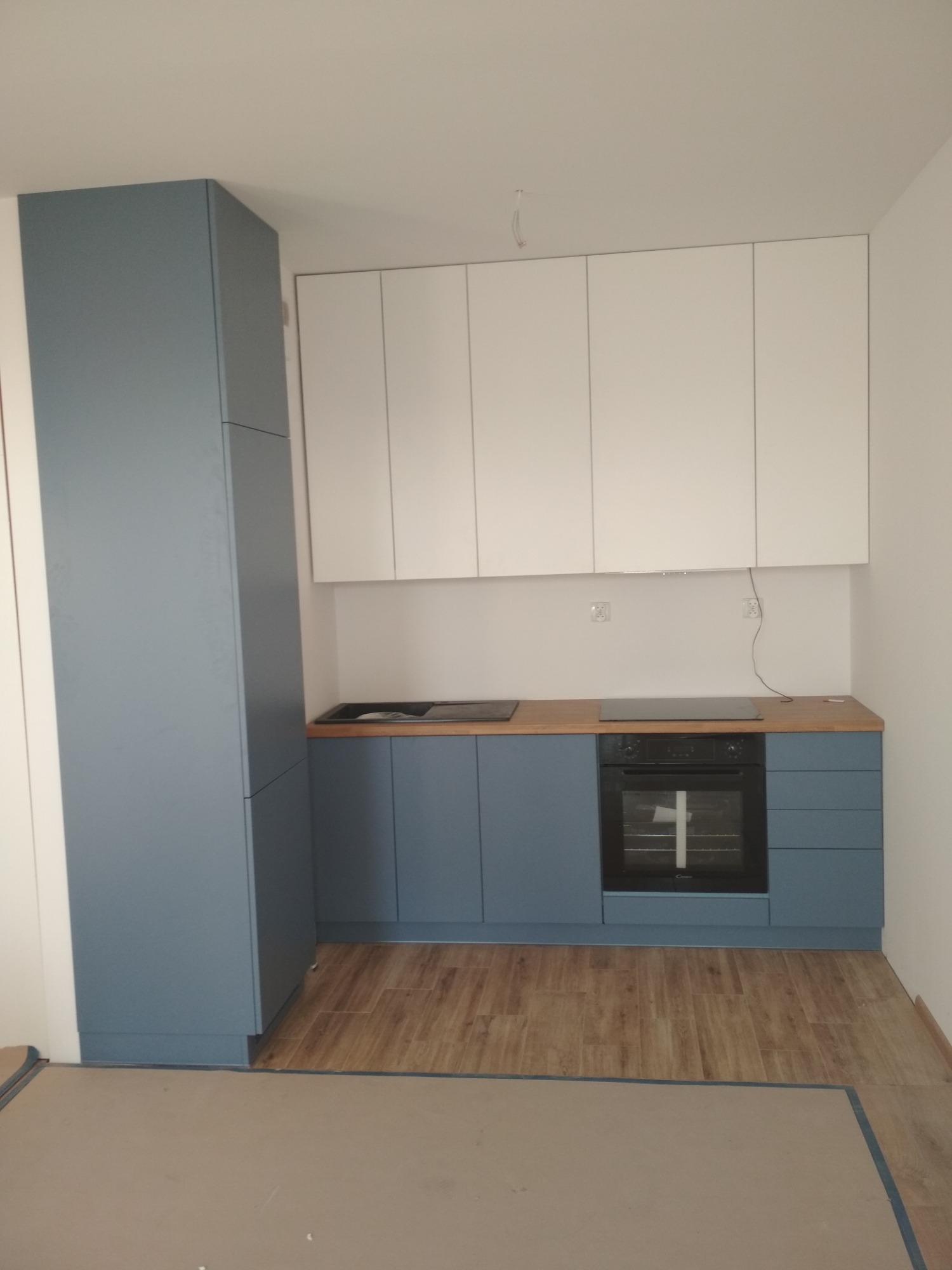 Nowoczesna kuchnia z niebieskimi szafkami dolnymi i białymi górnymi, drewniany blat, widok na płytę grzewczą i piekarnik, świeżo po montażu, z zabezpieczoną podłogą.