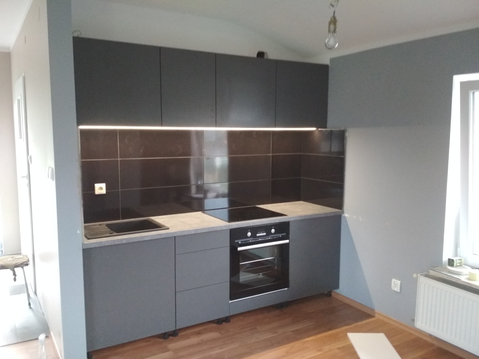 Nowoczesna, szara zabudowa kuchenna z górnymi szafkami podświetlonymi listwą LED, czarny panel nad blatem i sprzęt AGD w zabudowie, widok na fragment podłogi.