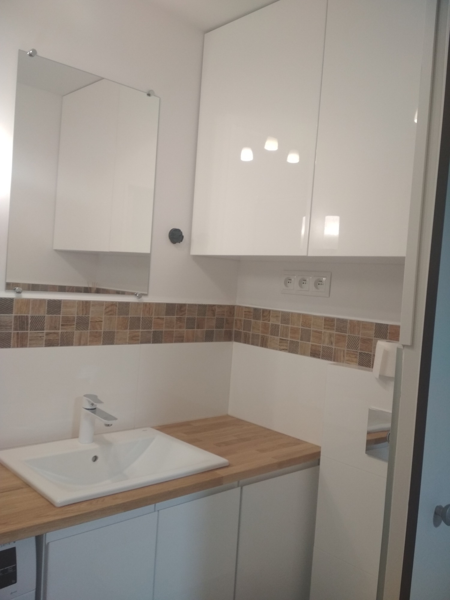 Nowoczesna łazienka w Toruniu z białą szafką pod umywalką z drewnianym blatem, lustrem i wiszącymi szafkami, wykończona mozaiką.