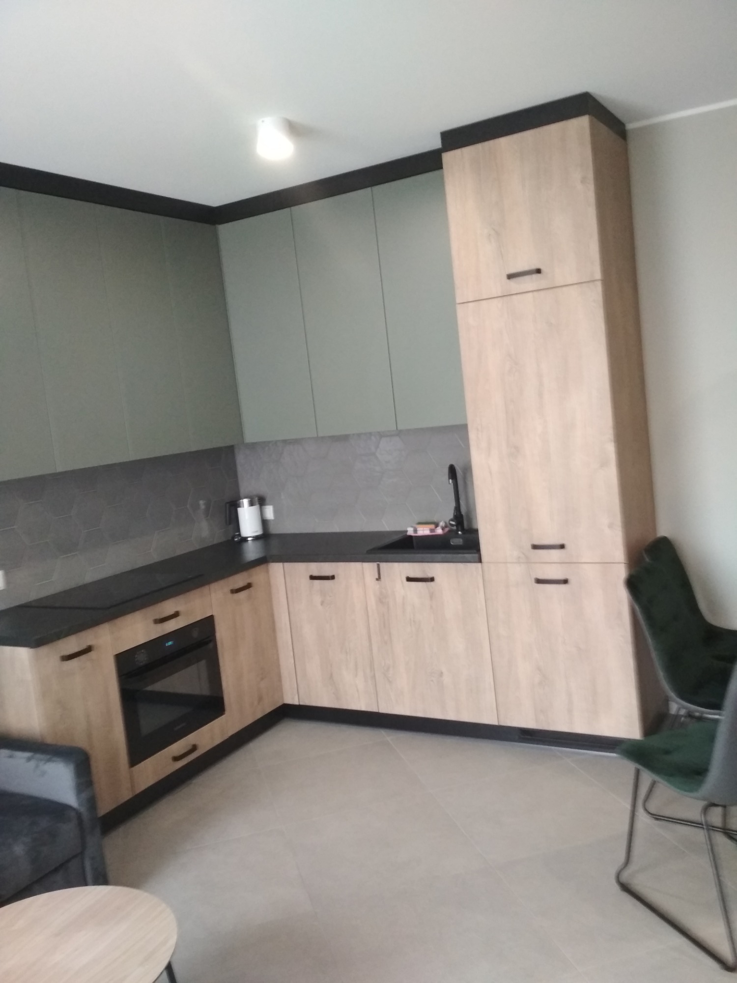 Nowoczesna kuchnia z drewnianymi szafkami i szarymi elementami, widok na zabudowę kuchenną z piekarnikiem i zlewem, heksagonalne płytki nad blatem, oświetlenie sufitowe.