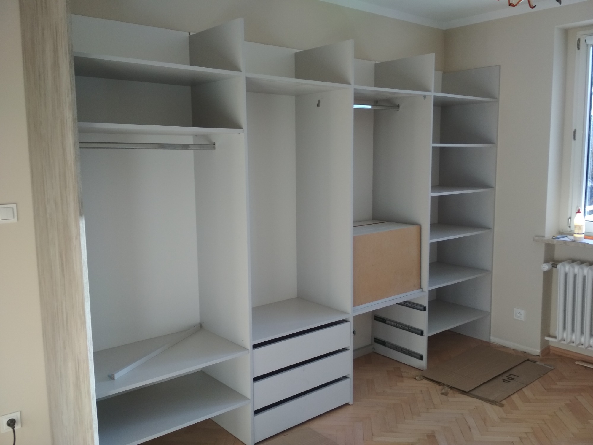 Wnętrze szafy wnękowej z białymi półkami i szufladami, widoczne prowadnice szuflad oraz karton na podłodze zabezpieczający przed zabrudzeniem.
