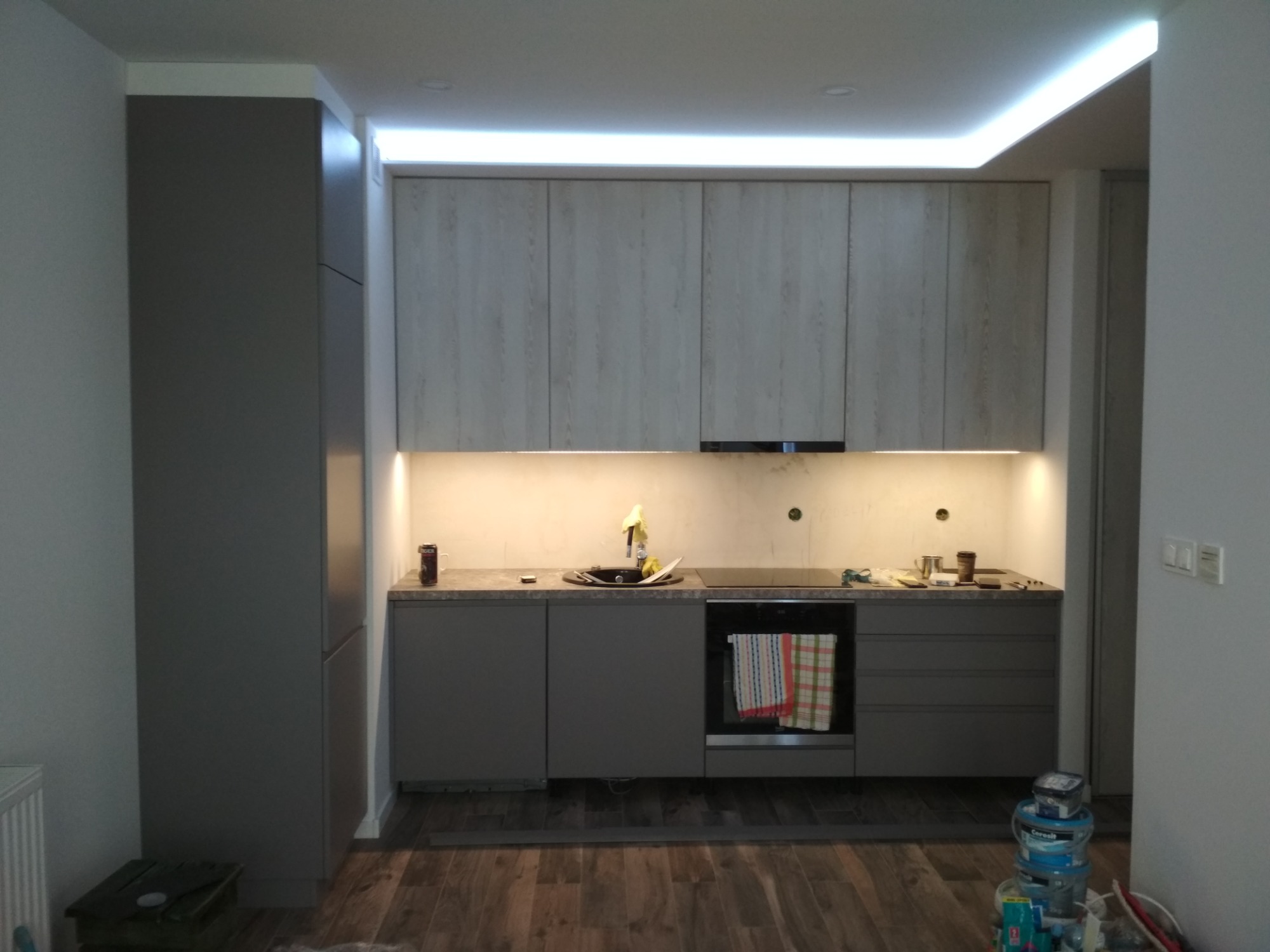 Szara zabudowa kuchenna z drewnopodobnymi szafkami wiszącymi i oświetleniem LED podszafkowym oraz sufitowym, widoczne niedokończone prace wykończeniowe i pojemniki z farbą na podłodze.